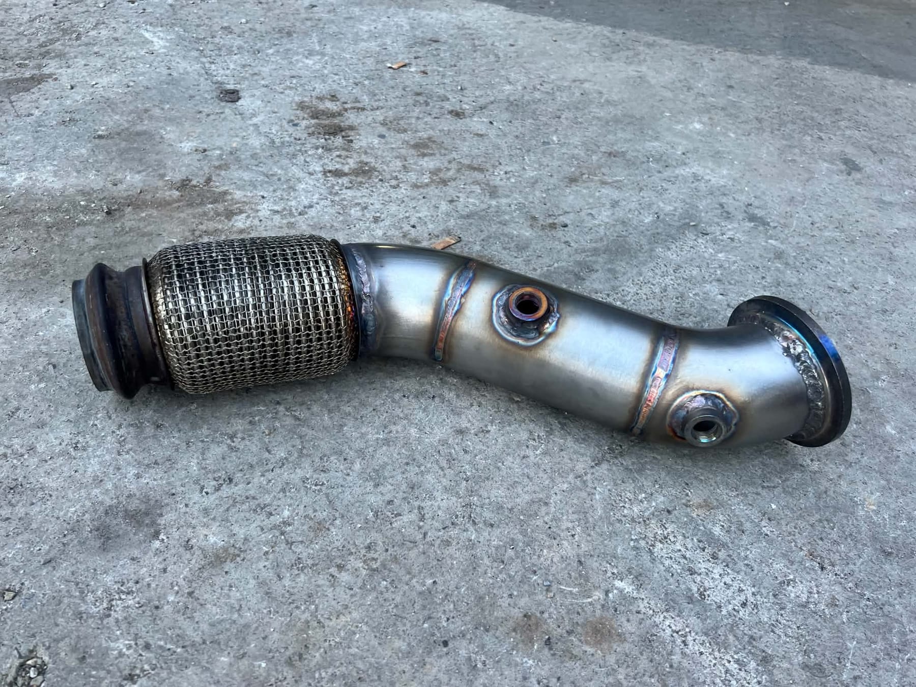 Një downpipe inoksi i përfunduar në dyshemenë e punëtorisë, me fleks të thurur, tegela të salduar me TIG dhe fole sensorësh me fileta.