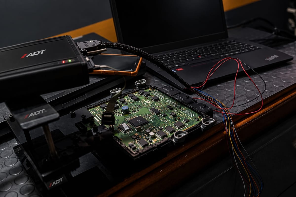 Njësia elektronike e motorit e hapur në bankun e punës, e lidhur me laptop dhe me pajisjen programuese, me vegla me shenjën ADT përreth.